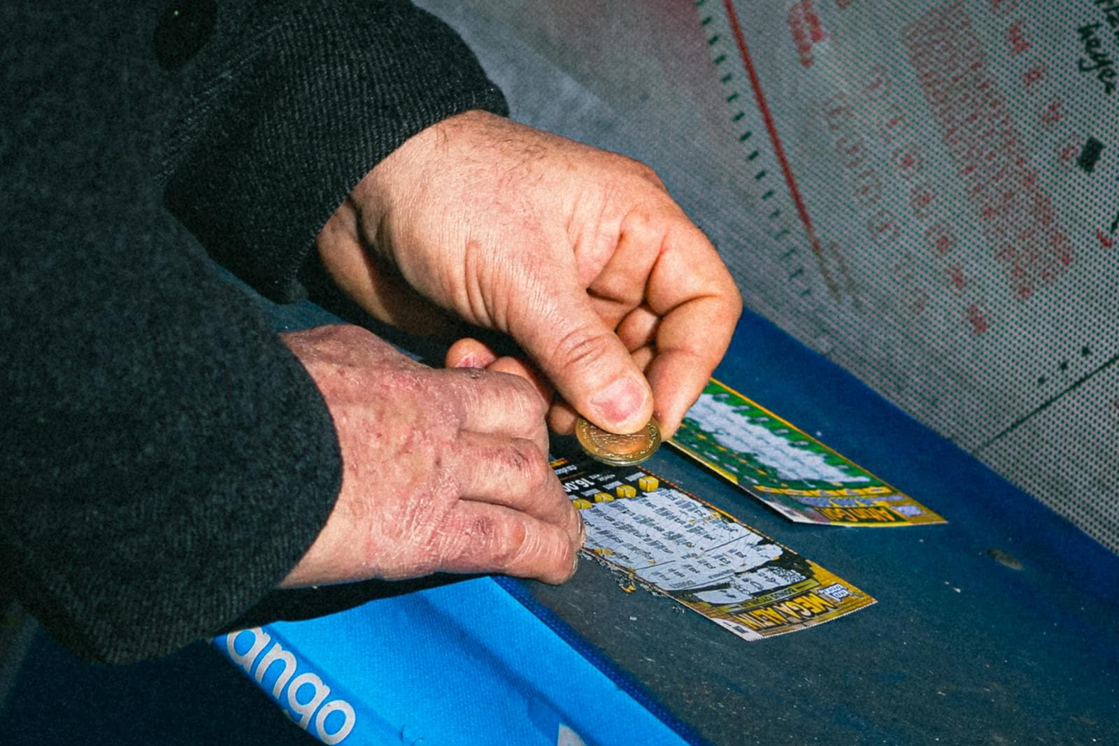 Hands using a coin to scratch off a lottery ticket on a blue surface.