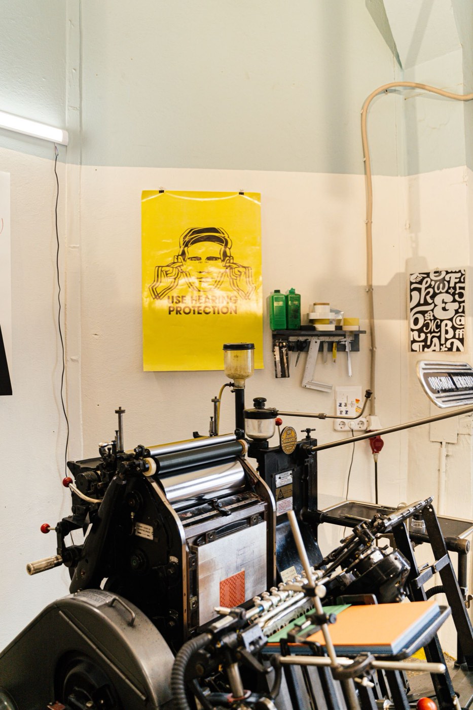 A printing press in a workshop with stacks of coloured paper stock beside it.